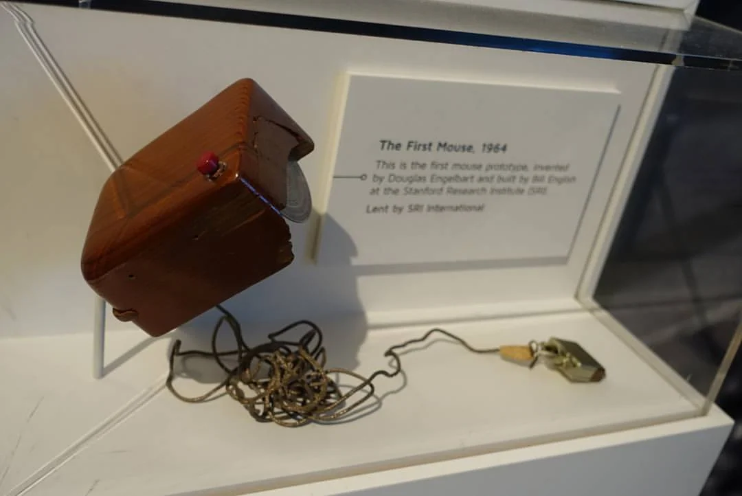 A museum case labelled The First Mouse, 1964, the wooden prototype tilted forward so its wheel is visible.