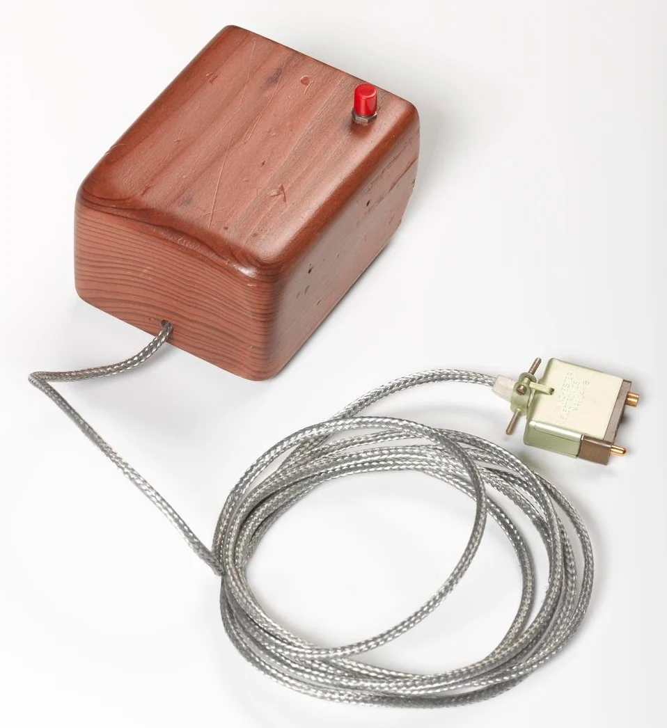 The first mouse photographed from above on white: a hand-sized block of shaped hardwood with a single red button and a braided steel cable.
