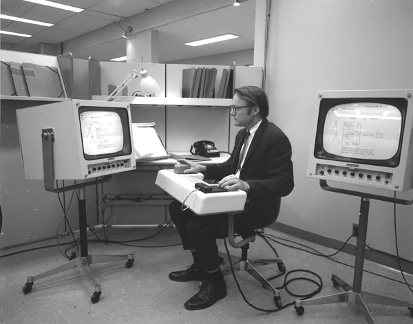 An NLS workstation in 1968: a researcher between two large cathode-ray tube displays, one hand on the mouse, the other on a five-key chord keyset.