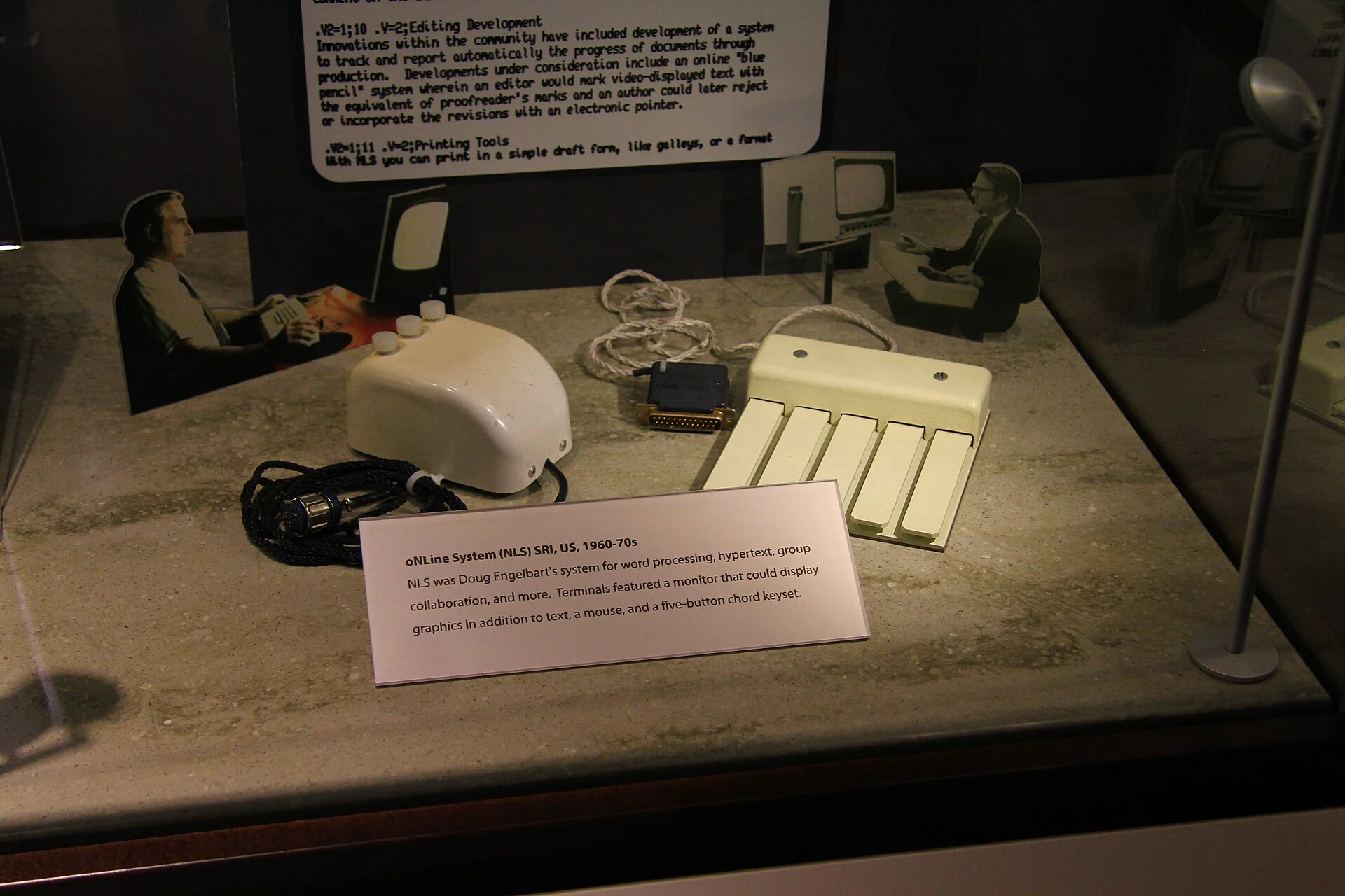 A three-button NLS mouse and a five-key chord keyset displayed together, with period photographs of NLS users mounted behind them.