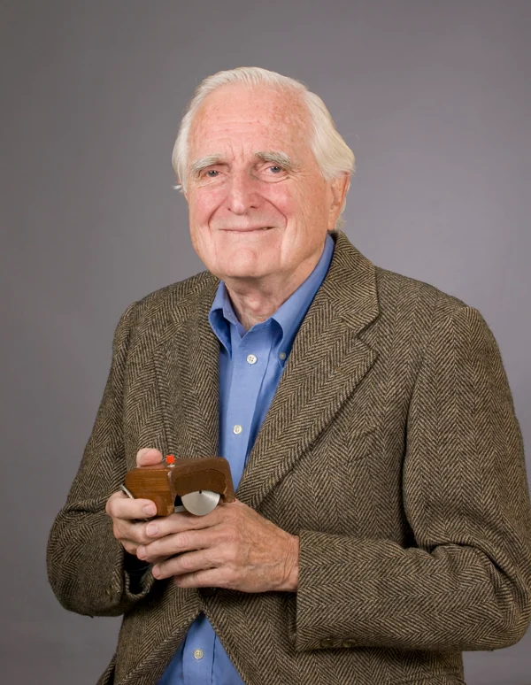 Douglas Engelbart in later life, holding the wooden prototype mouse in both hands and smiling at the camera.