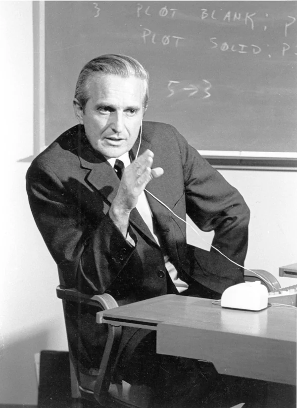 Douglas Engelbart in 1968, seated at a desk wearing a headset, one hand raised mid-sentence, a chalkboard of plotting commands behind him.