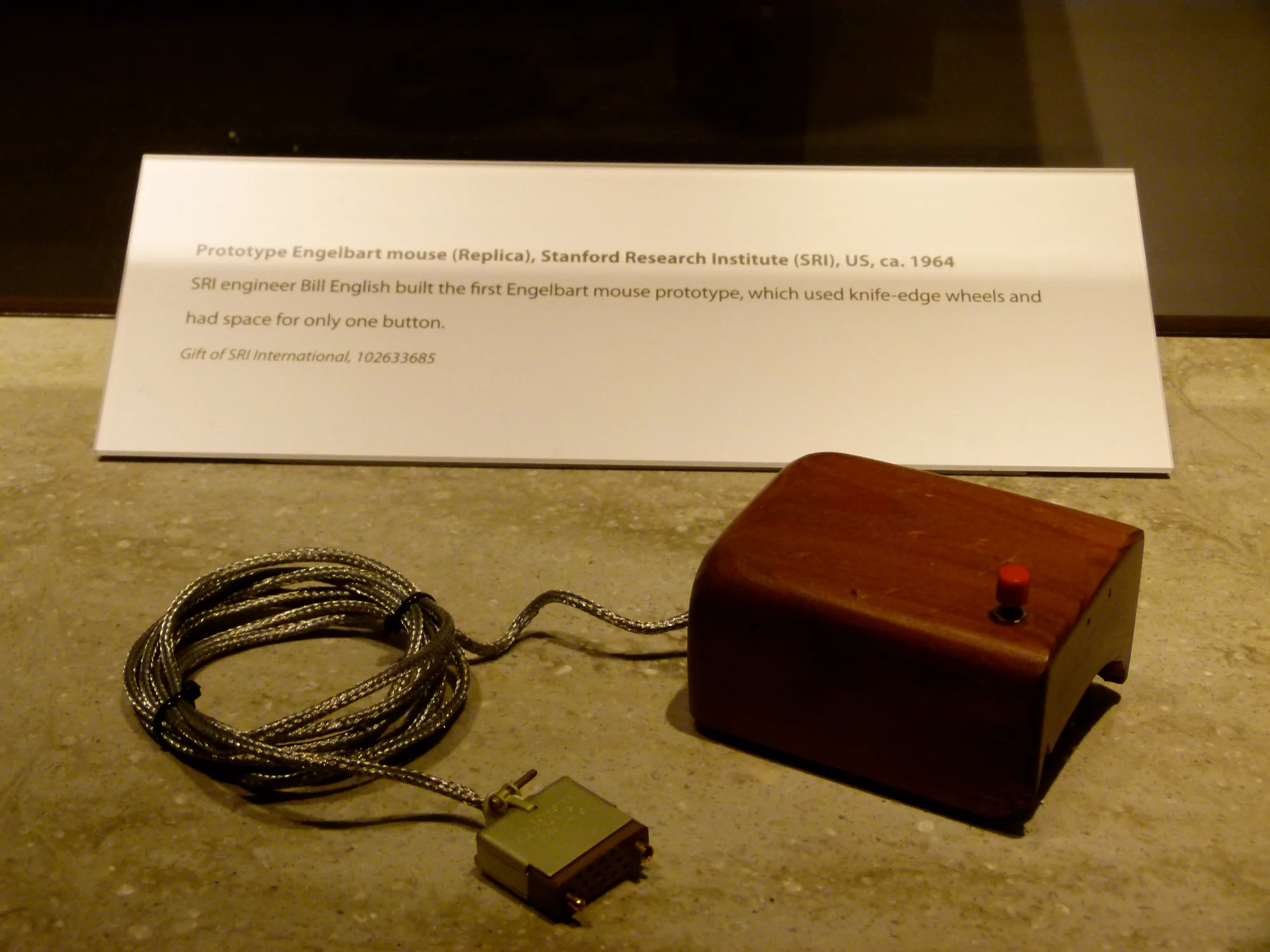 The prototype mouse in a museum vitrine beside its printed label, its cable coiled on the stone plinth.