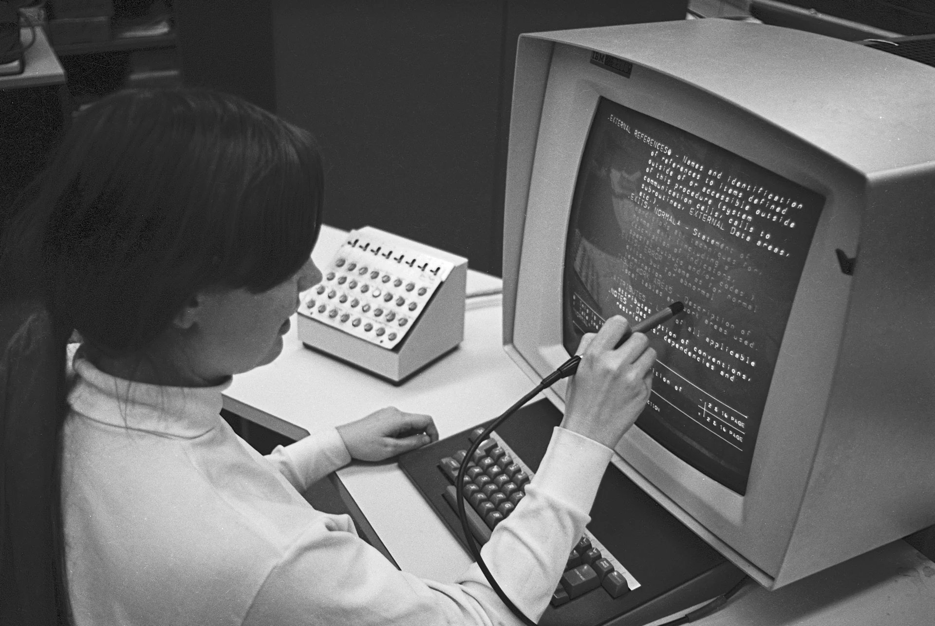 Over-the-shoulder view of a 1969 hypertext console, a light pen touching one line of text on a curved cathode-ray tube.
