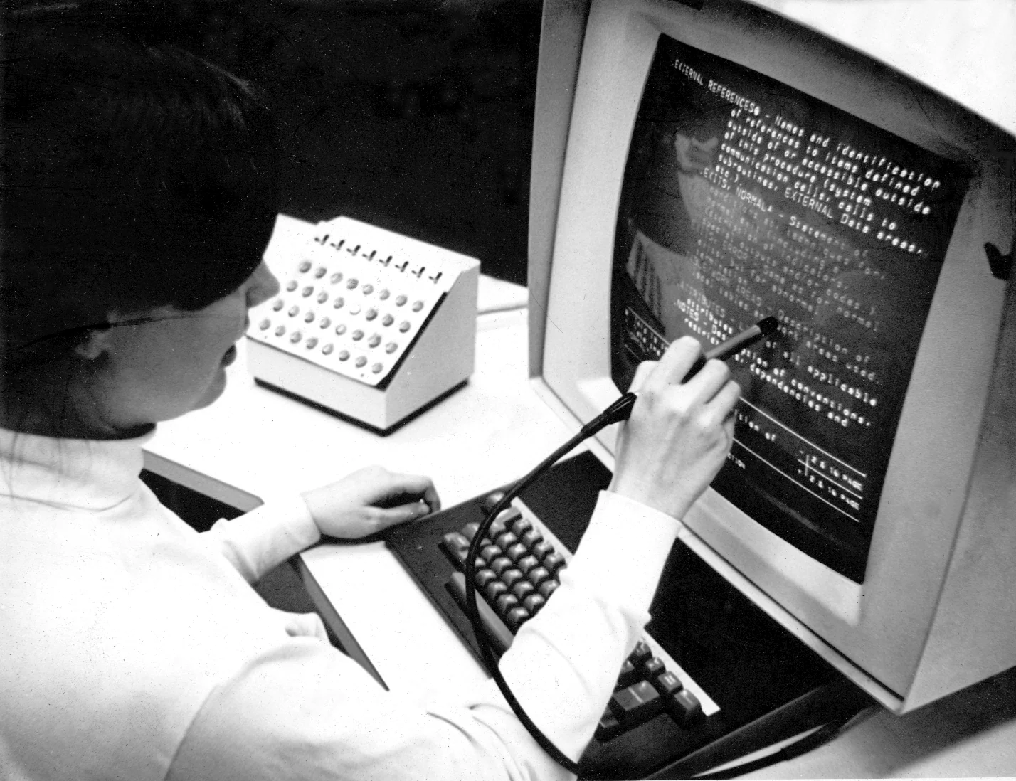 A woman at a 1969 display console reaching a light pen towards a screen of glowing text, a bank of round function buttons at her left.