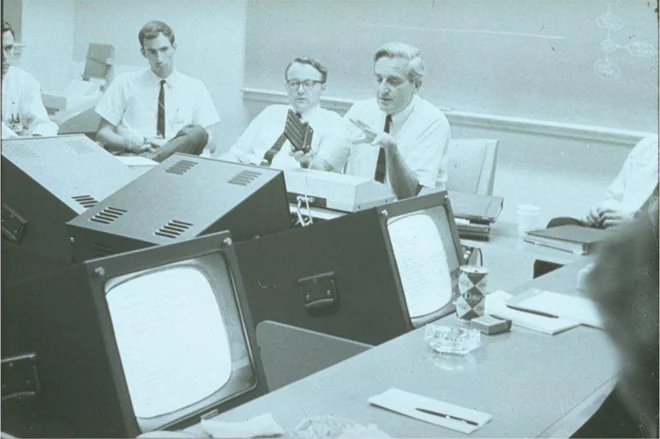 Researchers in shirtsleeves talking across a room lined with cathode-ray tube terminals showing pages of text.